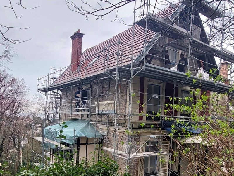 A house under renovation with scaffolding on all sides, workers present, and surrounded by trees.