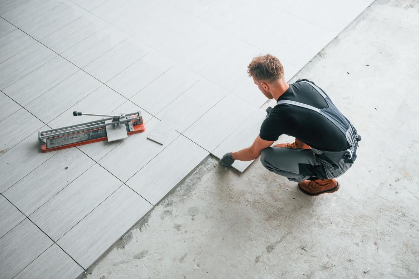 Man kneeling on floor installing gray tiles with a tile cutter beside him.